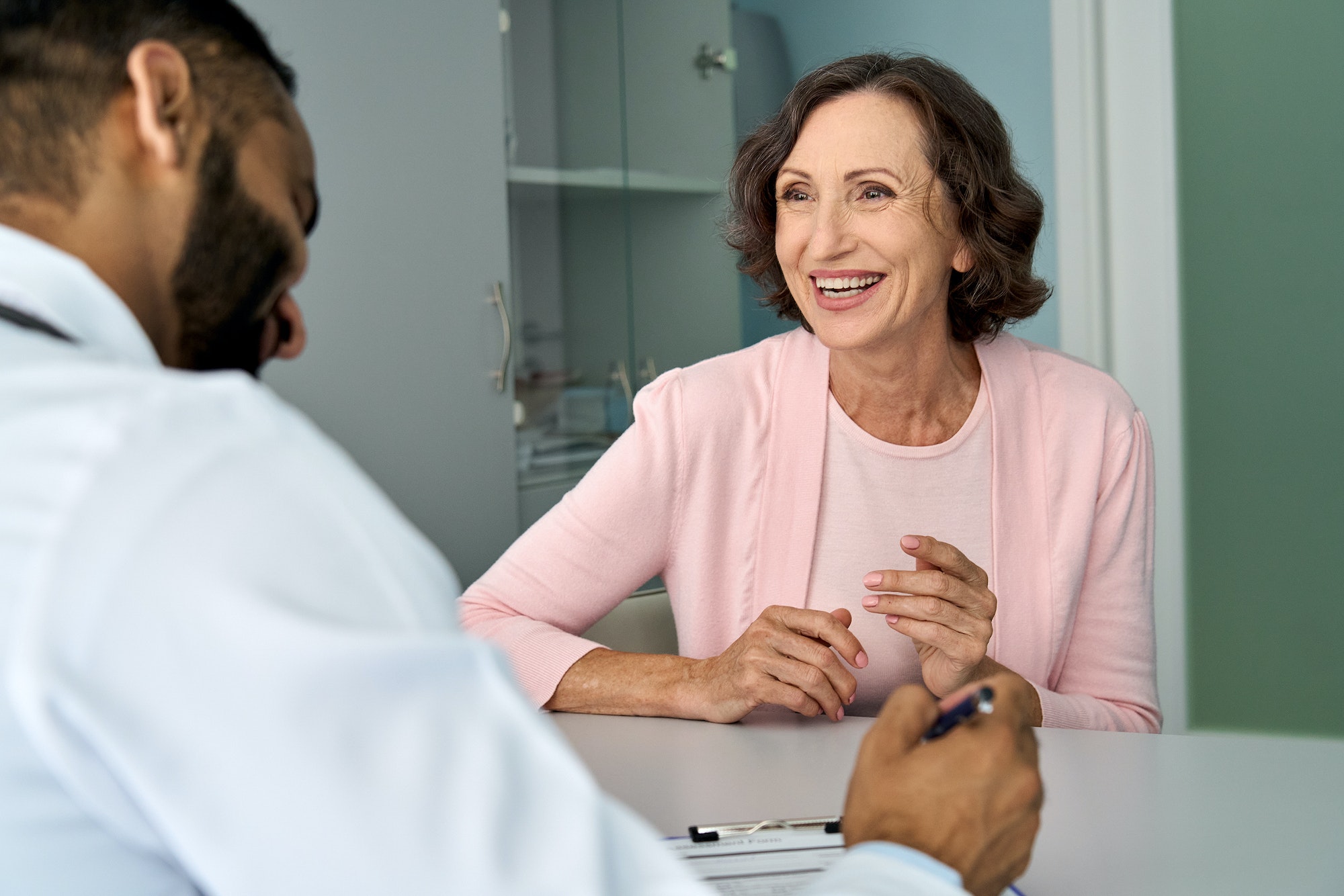 Happy older mature female patient talking to indian male doctor at appointment.